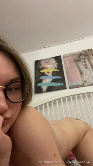 Woman with glasses, bare shoulders, sitting indoors near artwork.
