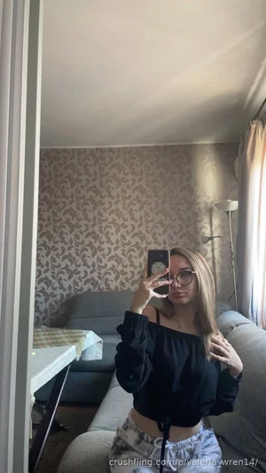 Woman with glasses in black off-shoulder top and patterned pants, sitting on couch in bedroom
