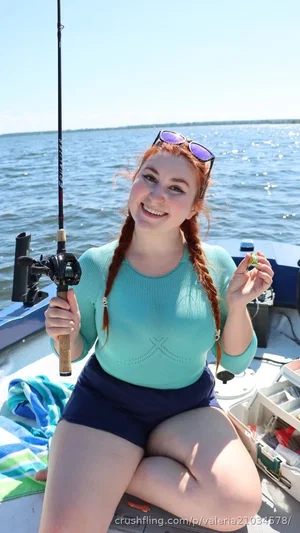 Woman with red braids in teal top and navy shorts, holding fishing rod on boat.
