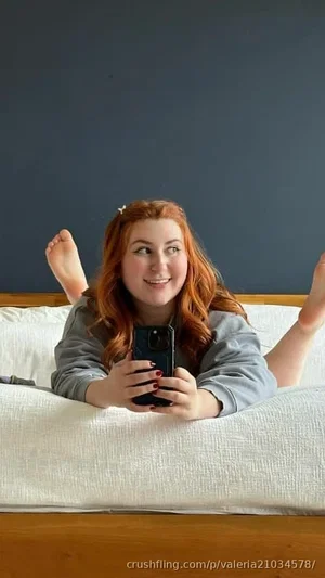 Woman with red hair in gray sweatshirt lying on bed, holding phone for selfie.