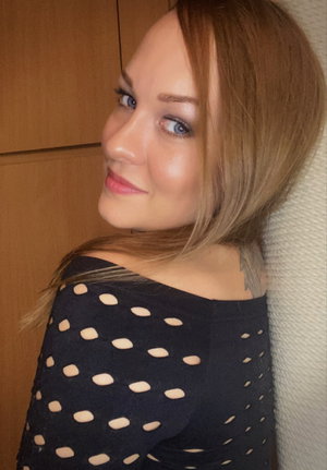 Woman with blonde hair in a black polka-dot top, posing indoors near a wooden cabinet.