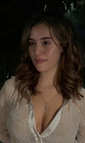 Woman with wavy brown hair in a white lace top, posing indoors at night.