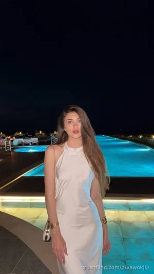 Woman in white halter dress standing by illuminated pool at night.