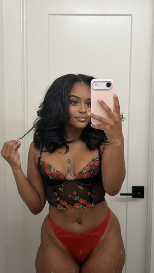 Woman in black floral lace top and red underwear poses in front of white door.