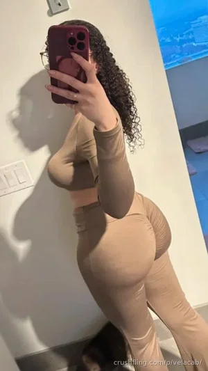 Woman with curly hair in beige long-sleeve top and matching pants, taking mirror selfie indoors.