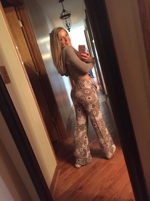 Woman with blonde hair in a long-sleeve crop top and patterned pants takes mirror selfie in hallway.