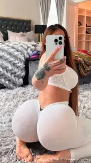 Woman with long hair and tattoos in white lace top and leggings, kneeling on rug in bedroom.