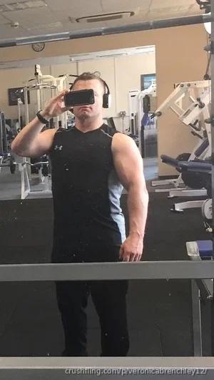 Man in black tank top and pants takes mirror selfie in gym.