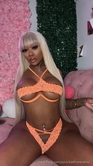 Woman with long blonde hair in orange lace lingerie sitting on pink furniture indoors.