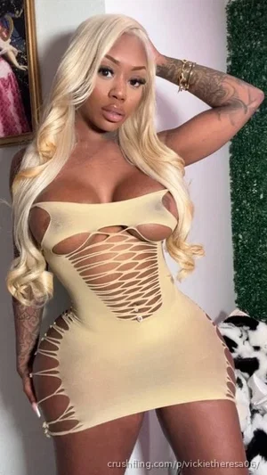 Woman with blonde wig, tattoos, wearing beige cutout dress, posing indoors.