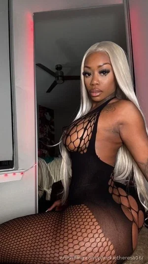 Woman with long blonde hair in black mesh bodysuit and fishnet stockings, posing indoors.