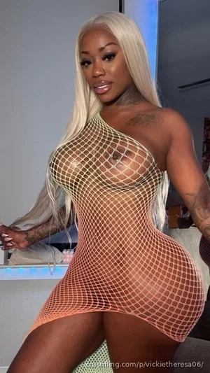 Woman with long blonde hair in a fishnet bodysuit posing indoors.
