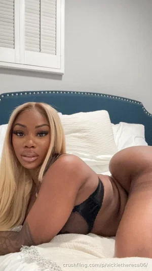 Woman with blonde hair in black lingerie lying on bed, indoor bedroom setting.