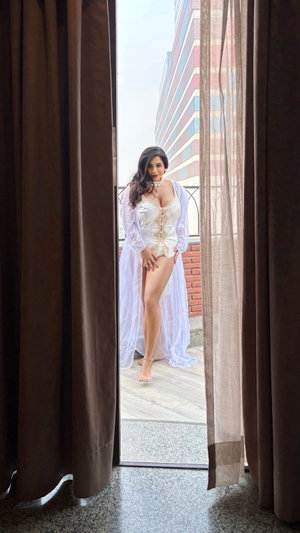 Woman in white lace lingerie and sheer robe standing on balcony, framed by curtains.