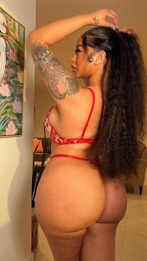Woman with long dark hair and tattoos wears red lingerie, standing indoors.