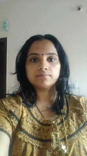 Dark-haired woman in yellow floral top with gold necklace indoors.