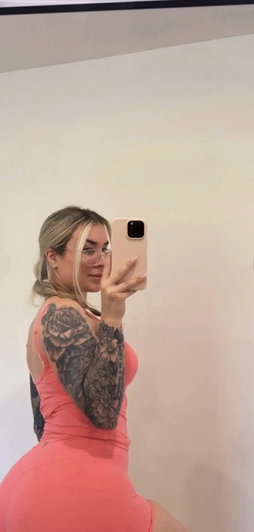 Woman with blonde hair and tattoos wears a pink dress, taking a mirror selfie indoors.