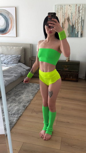 Woman in bright green and yellow outfit takes mirror selfie in bedroom.