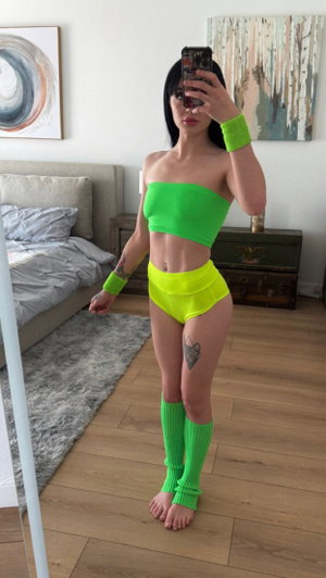 Woman in bright green and yellow outfit takes mirror selfie in bedroom.