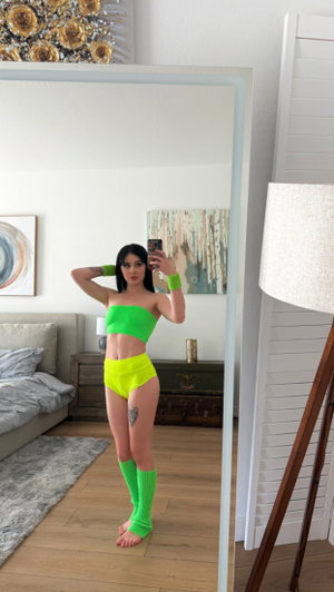 Woman in bright green and yellow outfit takes mirror selfie in bedroom.
