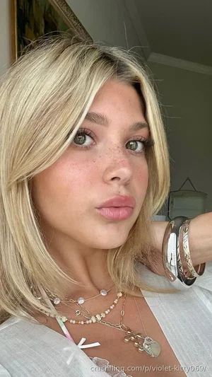 Blonde woman with freckles wearing layered necklaces and white top, indoor selfie.