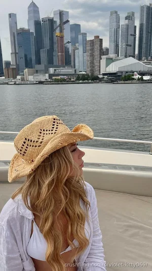 Woman with long blonde hair wearing straw hat and white top, sitting on boat with city skyline in