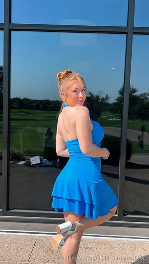 Woman in blue dress posing outdoors near glass wall, blonde hair in bun, wearing heels.