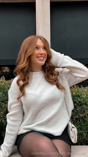 Woman with wavy red hair in white sweater and black skirt, sitting outdoors near greenery.