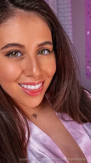 Woman with long brown hair in a light purple satin top, smiling in a bedroom setting.
