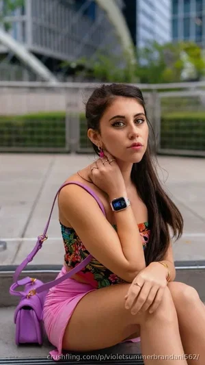 Woman with long dark hair in floral top and pink skirt, sitting outdoors near modern buildings.