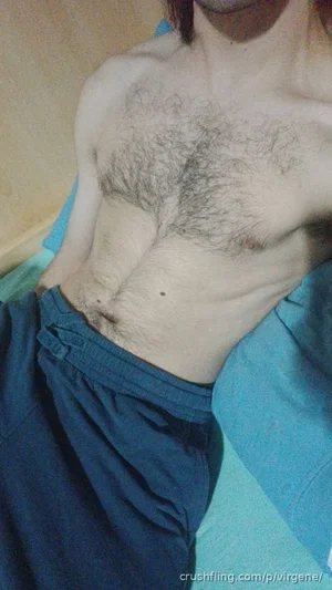 Shirtless man with chest hair lying on a bed, wearing blue pants.