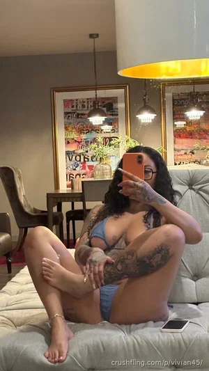 Woman with tattoos in blue lingerie sitting on couch, holding phone for selfie.