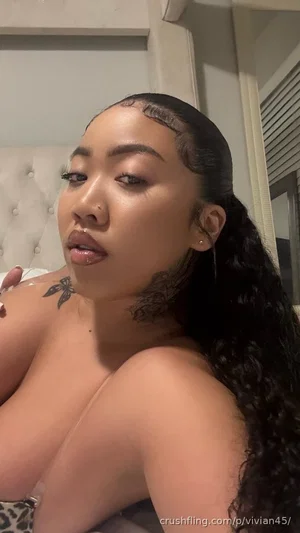 Woman with long dark hair and shoulder tattoo, topless, in bedroom setting.