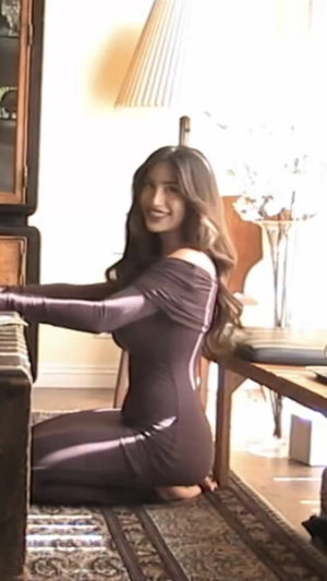 Woman in shiny brown bodysuit kneeling indoors near furniture, smiling.