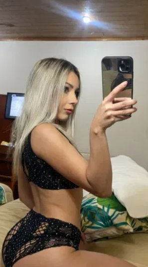 Blonde woman in sparkly black outfit taking mirror selfie in bedroom.