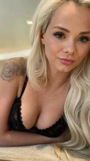 Blonde woman in black lace lingerie posing indoors, tattoo on shoulder.