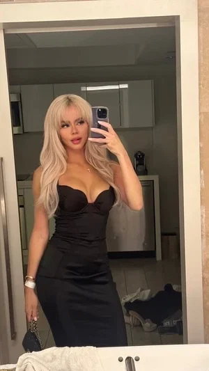 Woman with blonde hair in black dress taking mirror selfie in kitchen.
