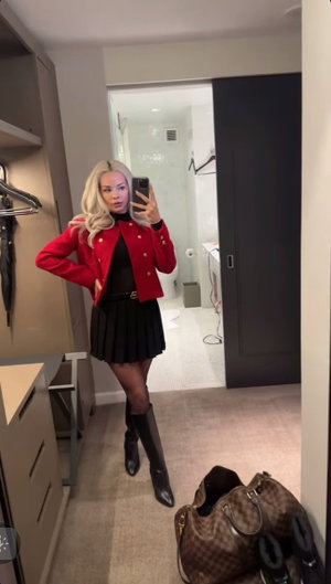 Blonde woman in red jacket, black dress, and boots poses in mirror selfie in bedroom.