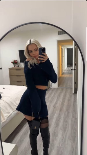 Woman with blonde hair in dark blue crop top and skirt, thigh-high boots, mirror selfie in bedroom.