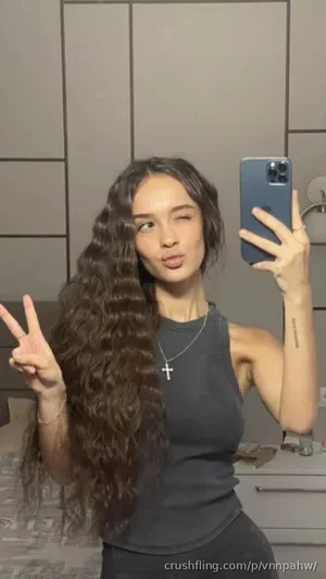 Woman with long curly hair in dark tank top takes mirror selfie in bedroom.