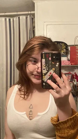 Woman with glasses and brown hair in white tank top takes mirror selfie in bedroom.
