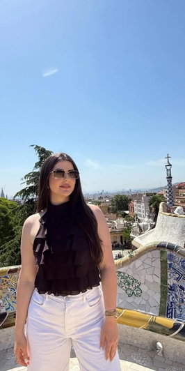 Woman with long dark hair in black top and white pants at outdoor viewpoint.