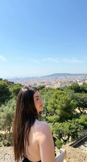 Woman with long dark hair in black top overlooking city from hill.