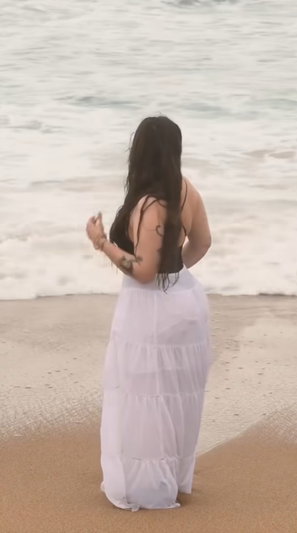 Woman with long dark hair in white skirt on beach.