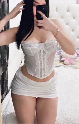 Woman with long black hair in white lace corset and skirt taking mirror selfie.