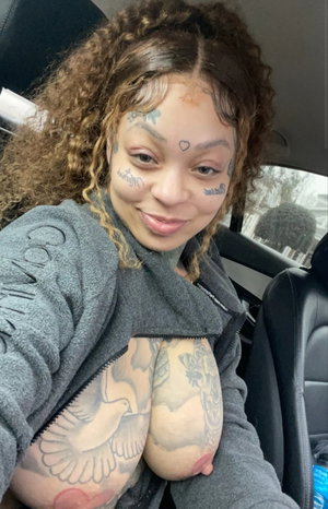 Woman with curly hair and tattoos in a car wearing gray jacket.