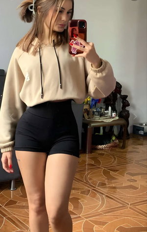 Woman in beige hoodie and black shorts taking indoor mirror selfie.