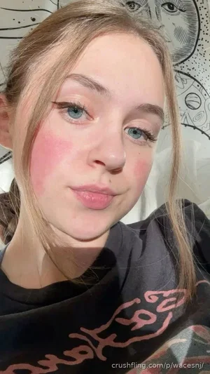 Woman with blonde hair and blue eyes wearing a black t-shirt, indoors with patterned background.