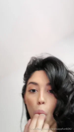 Woman with dark curly hair, applying lipstick, close-up face shot against plain background.