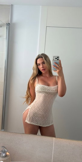 Woman with long blonde hair in a white lace bodysuit taking a mirror selfie in a bathroom.